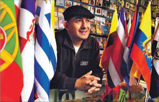  ?? MICHAEL STUPARYK/TORONTO STAR ?? Joe Nuñez looks out over a counter filled with South American flags at Super Latin Music on St. Clair Ave. W. The store also does a brisk business in Latin groceries, selling everything from cactus chunks to chili peppers. You can find about two-dozen...