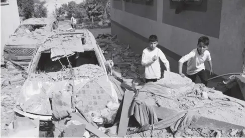  ?? ?? Two boys are seen standing next to a car that is heavily damaged and covered in debris in Gaza. In the foreground is what appears to be the rubble of a collapsed building.