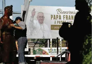  ?? José Luis Saavedra-10.jan.18/Reuters ?? Homem limpa escultura de mapuche diante de cartaz que dá as boas-vindas ao papa Francisco em Temuco, no Chile; de lá, pontífice viaja quinta para o Peru