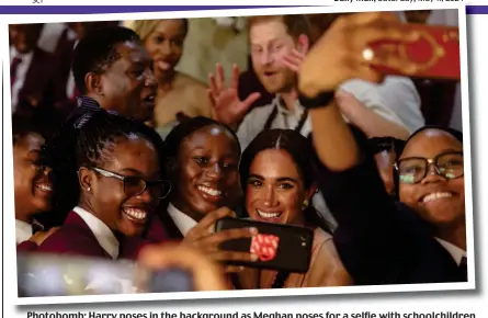  ?? ?? Photobomb: Harry poses in the background as Meghan poses for a selfie with schoolchil­dren