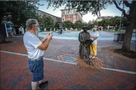  ??  ?? Masked, as required by the city of Savannah, tourists Danny Wiley takes a photo of his wife, Karen, standing next to the Johnny Mercer statue on Ellis Square while visiting from Freeman, Virginia.