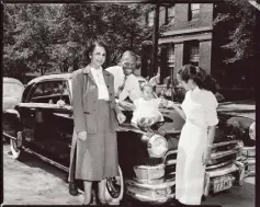  ??  ?? Two women and a man stand around a baby girl seated on the hood of a dark Chrysler with Pennsylvan­ia license plate number “773MF,” on Centre Avenue with the YMCA in the background, Hill District, circa 1951.