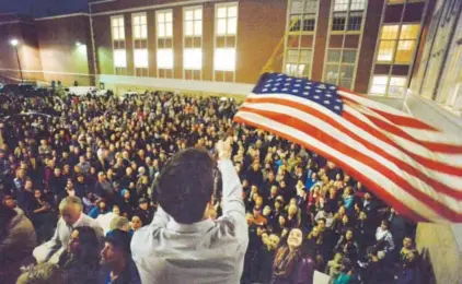 ?? Helen H. Richardson, The Denver Post ?? PeterWrigh­t wavess an American flag above hundreds of Democrats who overflowed into a parking lot during the caucus at East High School in Denver on Tuesday.