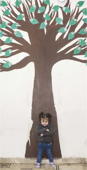  ??  ?? Three-year-old Nour, who was found alone and injured on the streets of Aleppo, stands in front of a tree decorated with the photos of other children who have been brought to a care facility for separated children. Photo: Mark Condren