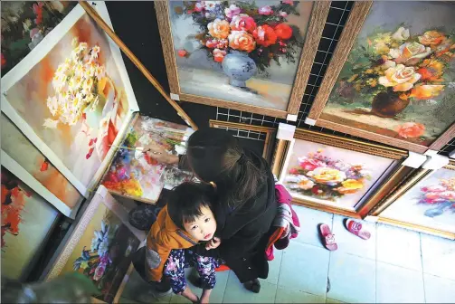  ?? XU YUANCHANG / FOR CHINA DAILY ?? An oil painter works on a still life painting as her daughter sits on her lap at an art gallery in Dafen village, Shenzhen, Guangdong province.