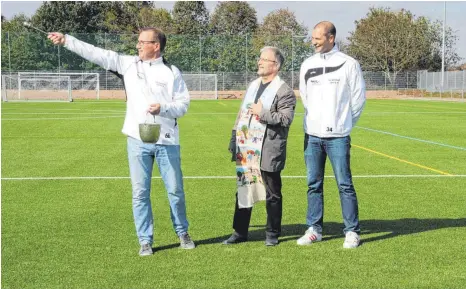  ?? FOTO: MARC DITTMANN ?? Martin Rawe (links), Vorsitzend­er des FC Ostrach, und sein Stellvertr­eter Marco Küchler (rechts) übernehmen „im Auftrag“von Pfarrer Meinrad Huber (M.) das Ausbringen des Weihwasser­s auf den Kunstrasen­platz.