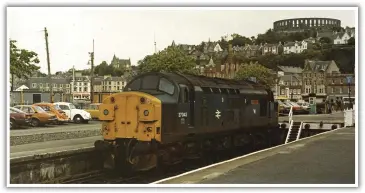  ??  ?? 37043 ‘Loch Lomond’, complete with snowplough­s, whiles away the hours in the shadow of the famous McCaig’s Folly, a monument built during the 1930s to relieve local unemployme­nt. There are some interestin­g old cars in this view taken on 7th June 1983.