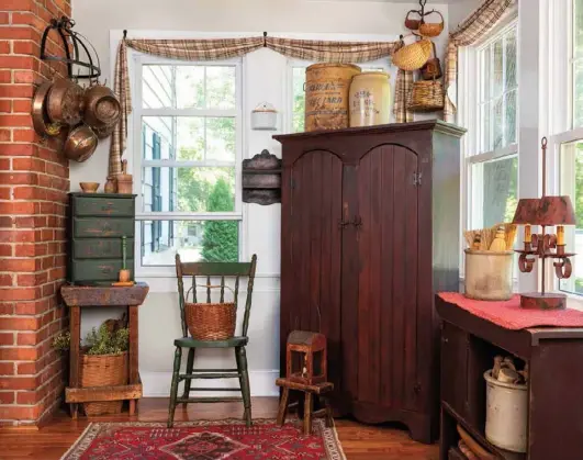  ??  ?? Kim anchors the kitchen gathering area with an oversize primitive cupboard and an Asian-style rug. She stacks and mingles functional pieces with decorative accents like the crock set inside the open cubby of a cabinet and the small chest of drawers perched on a wooden stand. To add visual balance to the setting, she mounted a pot rack hung with copperware in one upper corner and a rack holding an assortment of baskets in the opposite one.