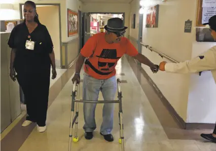  ?? Photos by Leah Millis / The Chronicle ?? Robert Hawkins gets a fist bump from lead cook Jonard Espinosa as nurse Hellane Merchant supervises his walk in Waters Edge skilled nursing home in Alameda. Hawkins was homeless before he was admitted to the nursing home.