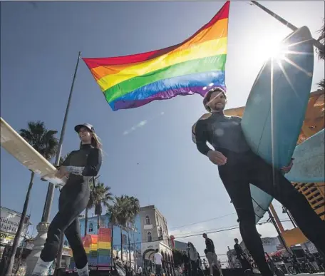  ?? Brian van der Brug Los Angeles Times ?? SURFERS head for the waves as a rainbow flag flies in Venice. The design of the first such banner is often solely credited to Gilbert Baker.