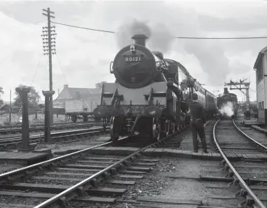  ?? M Carrier/Kiddermins­ter Railway Museum ?? Around 250 yards from Elgin station, on
24 May 1956 the signalman at Elgin East box reaches up with the train staff that will allow BR Standard ‘4MT’ 2-6-4T No 80121 to continue on the former Morayshire Railway route towards Lossie Junction, the fireman already leaning out for receipt. Lossie Junction is where the original Lossiemout­h line of August 1852 continues ahead and the Moray Coast route begins, turning east – the junction opened with the route to a temporary terminus at Garmouth on 12 August 1884, with completion of a ‘gap’ in the coast railway beyond there achieved on 5 April 1886. The line nearest the box, seemingly occupied by ex-CR 0-4-4T No 55221, is a lengthy headshunt, while that on the other side of the BR Standard is the route via Rothes to Craigellac­hie, with the connection to Elgin shed further over.