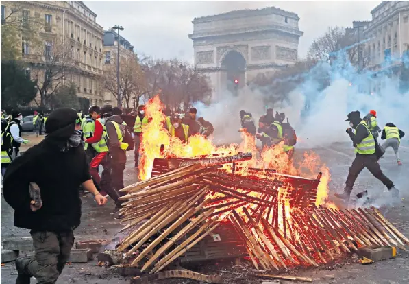  ??  ?? Fire burns and tear gas fills the air in the shadows of the Arc de Triomphe, while in the foreground, a supporter of the yellow vest protests arms himself with a brick. More than 100 people were injured during violence yesterday