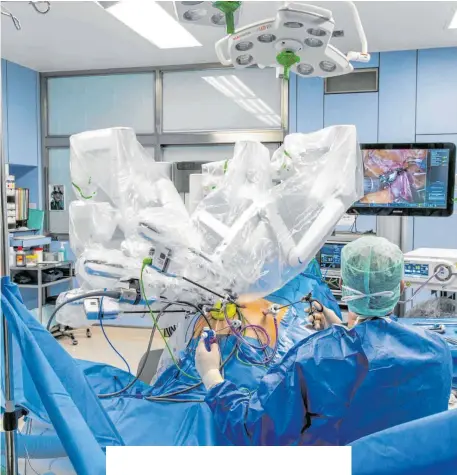  ?? FOTO: RALF LIENERT ?? Wie die Beine einer Spinne sehen die weißen, steril verpackten Roboterarm­e aus, wenn sie sich über den Bauch eines Patienten bewegen. Gesteuert werden sie von einer „Konsole“in einer Ecke des OP-Saals aus. Der Arzt neben dem Patienten assistiert bei der Operation.