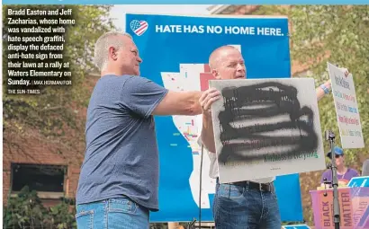  ?? | MAX HERMAN/ FOR THE SUN- TIMES ?? Bradd Easton and Jeff Zacharias, whose home was vandalized with hate speech graffiti, display the defaced anti- hate sign from their lawn at a rally at Waters Elementary on Sunday.