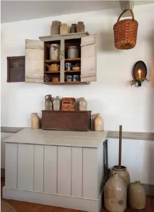  ??  ?? Below: A monochroma­tic color palette allows larger pieces, such as this firewood storage bin, to blend into the dining room without overtaking the space. An antique stoneware butter churn and jugs featuring similar coloration cluster beside the bin; smaller stoneware pieces are arranged in a hanging cupboard above.