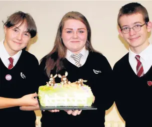  ??  ?? Zara Pugh, centre, with head girl Molly Cross and head boy Edward Ready at the Fearns Macmillan Coffee morning, holding the cake she and her grandmothe­r made which was star prize in the raffle