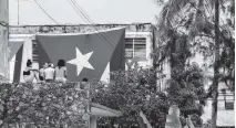  ?? AP ?? Neighbors look at Cuban flags that were draped over the windows of opposition activist Yunior Garcia’s home in an attempt to stop him from communicat­ing with the outside world during planned protests.