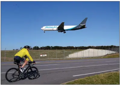  ?? (AP/Julio Cortez) ?? A cyclist rides nearby as an Amazon Prime jet from Miami Internatio­nal Airport makes its landing approach Monday at the airport in Glen Burnie, Md. Amazon is paying bonuses to employees this year to make sure there are enough people to handle the Christmas shipping rush.