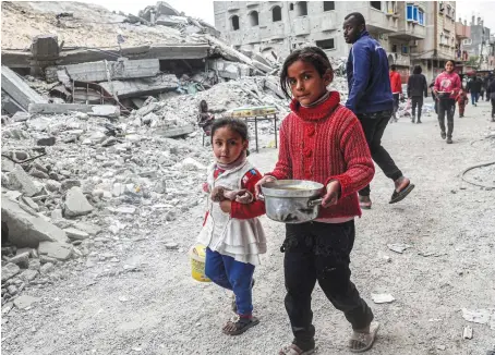  ?? AFP ?? Children walk past the rubble of a collapsed building with a pot of food provided by a charity organizati­on in Rafah in the southern Gaza Strip on Saturday.