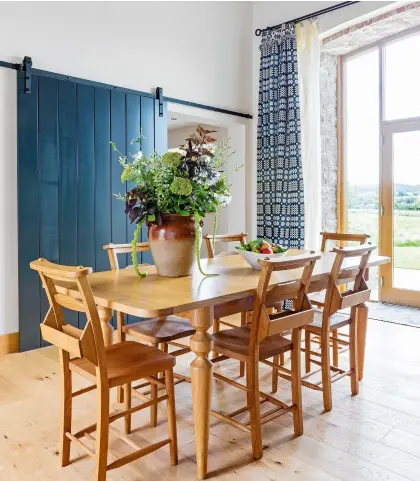  ??  ?? Left: A dining table from Made makes the most of the beautiful views through the full-height glass doors. Hanging from a rail made by nearby Celtic Dreams Ironwork, are traditiona­l Welsh weave curtains from Trefriw Woollen Mills. Zoë had to persuade them to leave the yarn on the loom to create the longer length needed. The new chapel chairs are from Peppermill Interiors
Opposite: The kitchen island is topped with a fine slab of slate, a relic from the barn’s previous life when it divided the cow stalls for milking. In the slate, probably mined nearby, are fossils adding a layer of prehistory to the 450-year-old barn. Above the Rangemaste­r oven is a splashback from Colour2gla­ss. The engineered oak flooring is from RM Jones Joinery in Ruthin