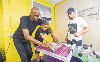  ?? KENNETH K. LAM/BALTIMORE SUN ?? Artist Mark Bradford, left, helps Ayden Brooks, 7, silk screen a T-shirt as Nicholas Mitchel, co-founder of Noisy Tenants, looks on at Greenmount West Power Press. The community workshop teaches kids how to do silk screening.