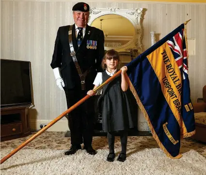  ??  ?? Mollie Stonelake, seven, holds a full-size standard from the Torpoint &amp; District Branch of the Royal British Legion, alongside branch chairman Colin Prideaux, in her home in Torpoint, Cornwall. Mollie will carry a specially-made miniature standard on Sunday in Torpoint