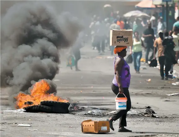  ??  ?? Une Haïtienne transporta­nt des provisions marche près d’un feu de pneus allumé par des manifestan­ts, hier, à Port-au-prince.