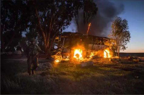  ?? Tsafrir Abayov/Associated Press ?? An Israeli soldier stands near a burning bus after it was hit by a mortar shell fired from Gaza near the Israel Gaza border Monday. Israel’s military says it will step up its efforts against Palestinia­n militants if rocket fire at Israel continues.