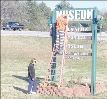  ?? Keith Bryant/The Weekly Vista ?? Xyta Lucas, left, president of the Bella Vista Historical Society, keeps an eye on the ladder while Benton County inmate Doug Headrick deals with peeling paint on the Bella Vista Historical Museum’s sign.