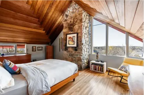  ?? ?? FROM THE TOP The living room features a waisthigh stone wall, as well as long, padded bench seating and a wall of windows; the primary suite offers exposed stone, hardwood flooring, and a cathedral ceiling clad in redwood planks; the home sits on more than an acre and does not have a garage; and the kitchen comes with a two-tier island that has one level of granite and one of glass.