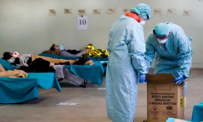  ??  ?? ITALIAN EXPERIENCE: Paramedics carry a hazardous medical waste box as patients lie on camping beds in an emergency structures set up at the Brescia hospital, Italy. Photo: Luca Bruno