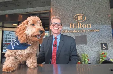  ?? MICHAEL SEARS / MILWAUKEE JOURNAL SENTINEL ?? Millie, the “canine concierge” at Hilton Milwaukee City Center, greets guests with her owner, Rusty Dahler, the hotel’s concierge. The dog already has a following on social media. For more photos and a video, go to jsonline.com/greensheet.