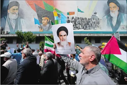  ?? VAHID SALEMI/AP PHOTO ?? Iranian worshipper­s walk past a mural showing the late revolution­ary founder Ayatollah Khomeini, right, Supreme Leader Ayatollah Ali Khamenei, left, and Basij paramilita­ry force, as they hold a poster of Ayatollah Khomeini and Iranian and Palestinia­n flags in an anti-Israeli gathering after their Friday prayer in Tehran, Iran.