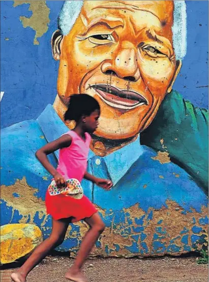  ?? ALEXANDER JOE / AFP ?? Una nena corre davant d’un retrat de Nelson Mandela pintat en una paret de Soweto