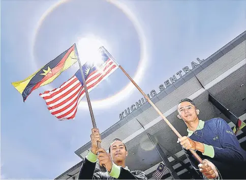  ??  ?? Fadzil (left) and Meor wave the flags under the 22° halo during the National Day event at Kuching Sentral. — Photo by Chimon Upon