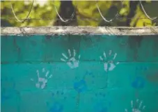  ??  ?? Kathleen Greeson juxtaposes the innocence of children’s handprints on a school wall with barbed wire topping the wall.