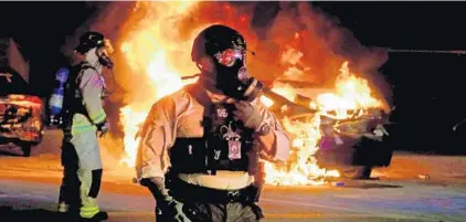  ?? MIKE STOCKER/SOUTH FLORIDA SUN SENTINEL ?? A police officer stands guard as firefighte­rs try to put out a fire in a burning car in downtown in Miami on Saturday as protests in the wake of the police killing of George Floyd turned destructiv­e.