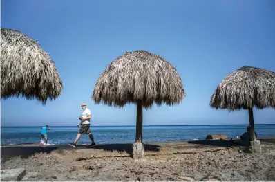  ?? Associated Press ?? A tourist, wearing a protective face mask amid the coronaviru­s pandemic, walks along the beach shore Tuesday in Havana, Cuba. The Caribbean is hunting for visitors and vaccines to jump-start the stalled economy in one of the world’s most tourism-dependent regions.