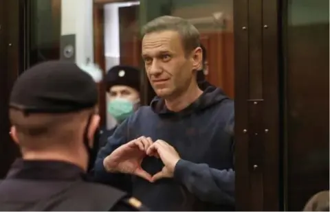  ?? FOTO VIA REUTERS ?? Terwijl de rechter het vonnis voorleest, maakt een grijnzende Navalny een hartje naar zijn vrouw Joelia.