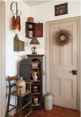  ??  ?? Right: Filling an empty corner or cubby with angled pieces, like this slender cupboard and miniature hanging shelf, introduces primitive appeal into what might otherwise be dead space in a kitchen corner. Similar-sized stoneware and pottery on the shelves convey a streamline­d aesthetic, punctuated by a vintage high chair, hanging drip candles, and a twig-and-berry wreath on the adjacent door. 45