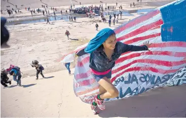  ?? RAMON ESPINOSA/ASSOCIATED PRESS ?? A migrant woman helps carry a handmade U.S. flag up the riverbank at the Mexico-U.S. border after getting past Mexican police at the Chaparral border crossing in Tijuana, Mexico, on Sunday.