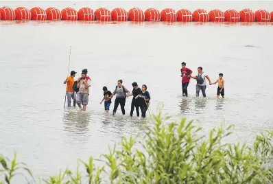  ?? (AP photo/ Eric Gay) ?? Migrants who crossed the Rio Grande from Mexico walk past large buoys being deployed as a border barrier on the river July 12 in Eagle Pass, Texas. The floating barrier is being deployed in an effort to block migrants from entering Texas from Mexico.