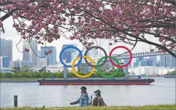  ?? (AP/Eugene Hoshiko) ?? Under blooming cherry blossoms, people wearing protective masks to help curb the spread of the coronaviru­s walk with a backdrop of the Olympic rings floating in the water in the Odaiba section of Tokyo.