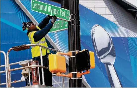  ?? CURTIS COMPTON / CCOMPTON@AJC.COM ?? Department of Public Works employee Deontie Parker installs a new street sign on Centennial Olympic Park Drive NW in preparatio­n for the Super Bowl with Mercedes-Benz Stadium behind him Thursday.
