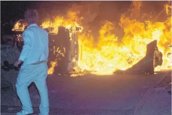  ?? FOTO: DPA ?? Vor einem brennenden Pkw auf einer Straße am zentralen Asylbewerb­erheim in Rostock-Lichtenhag­en: Die Bilder im Sommer 1992 erschütter­ten die Republik.